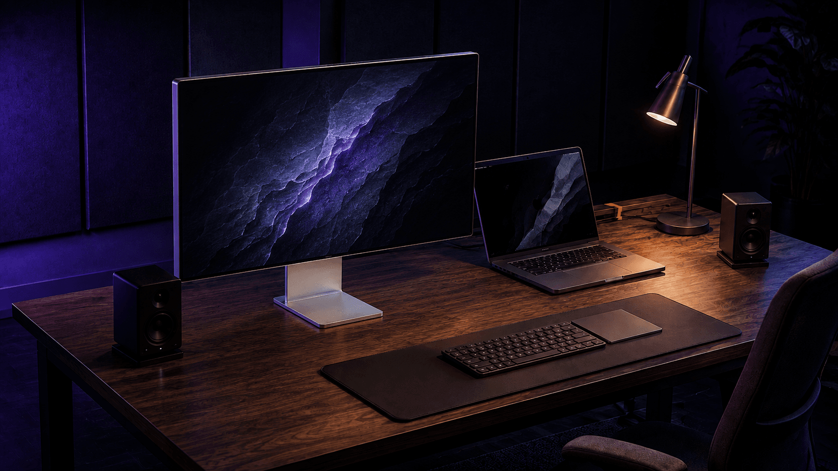 Estudio digital conceptual con monitor de frente, portátil abierto a la derecha, teclado, trackpad, dos altavoces y lámpara sobre una mesa de madera oscura.